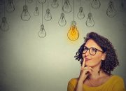 Portrait thinking woman in glasses looking up with light idea bulb above head isolated on gray wall background